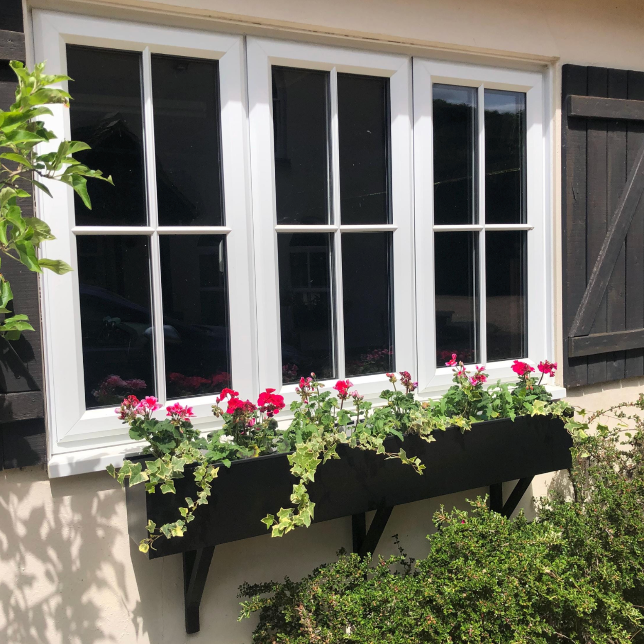 Window with black shutters and a flower box filled with flowers on a white wall.