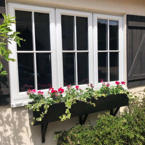 Window with black shutters and a flower box filled with flowers on a white wall.